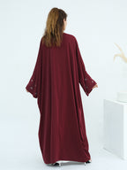 A woman viewed from behind wearing a long, flowing abaya dress in deep burgundy with embroidered lace cuffs, standing against a clean white studio background.