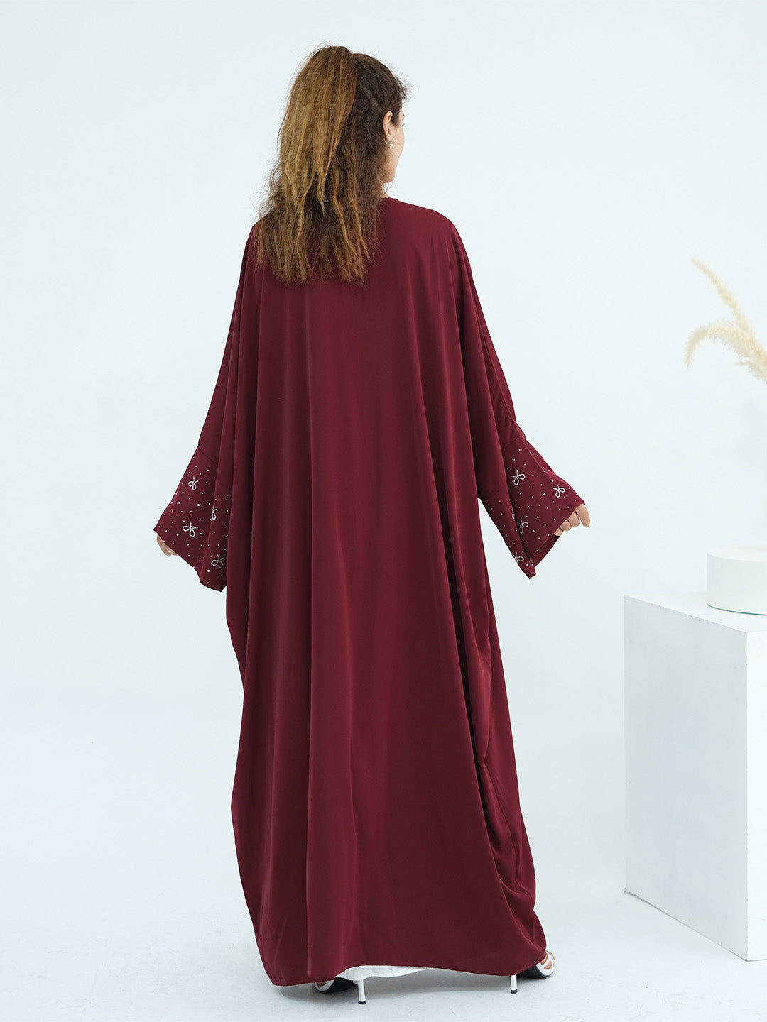 A woman viewed from behind wearing a long, flowing abaya dress in deep burgundy with embroidered lace cuffs, standing against a clean white studio background.