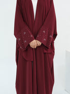 A person wearing a loose, flowing burgundy abaya with wide sleeves and decorative silver bow and dot embroidery on the cuffs, shown against a plain white background.