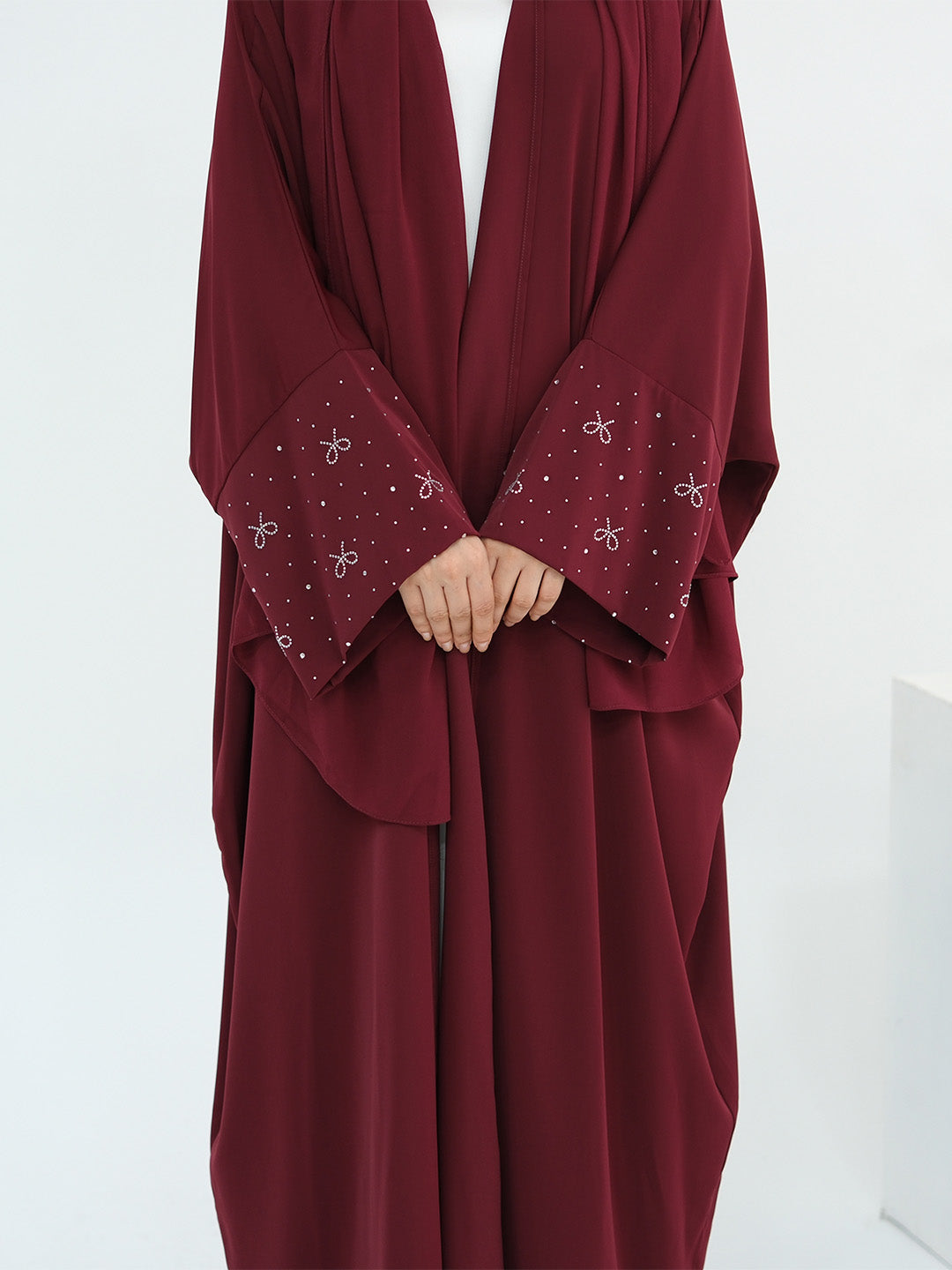 A person wearing a loose, flowing burgundy abaya with wide sleeves and decorative silver bow and dot embroidery on the cuffs, shown against a plain white background.