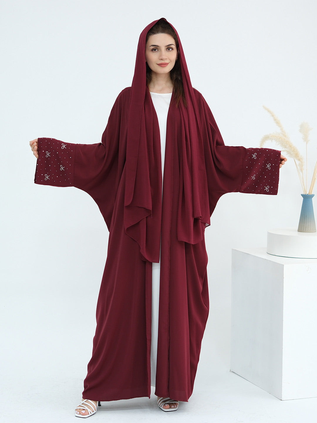 A woman modeling a deep burgundy hooded abaya with wide draped sleeves and embroidered cuffs, worn over a white inner dress. She is standing in a bright studio setting with a white pedestal and dried pampas grass in a vase.