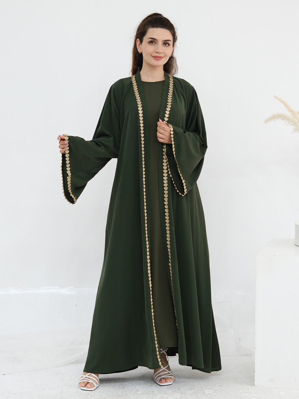 A woman modeling a dark green abaya dress with wide bell sleeves and gold chain trim along the front opening, shown against a clean white studio background.