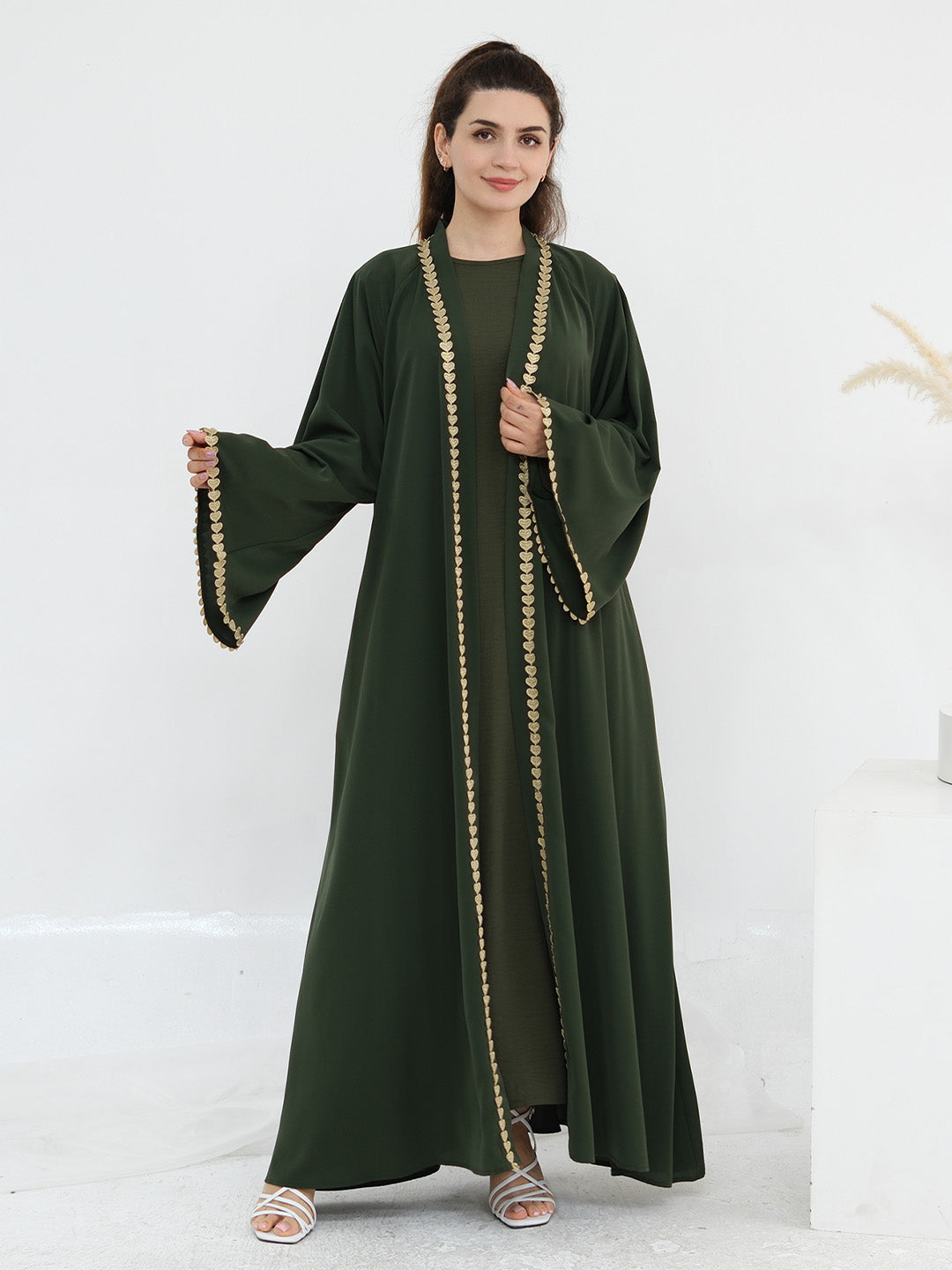 A woman modeling a dark green abaya dress with wide bell sleeves and gold chain trim along the front opening, shown against a clean white studio background.