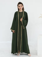 A woman modeling a dark green abaya dress with gold beaded trim down the front and along the sleeve cuffs, paired with a matching green inner dress, against a clean white studio background.
