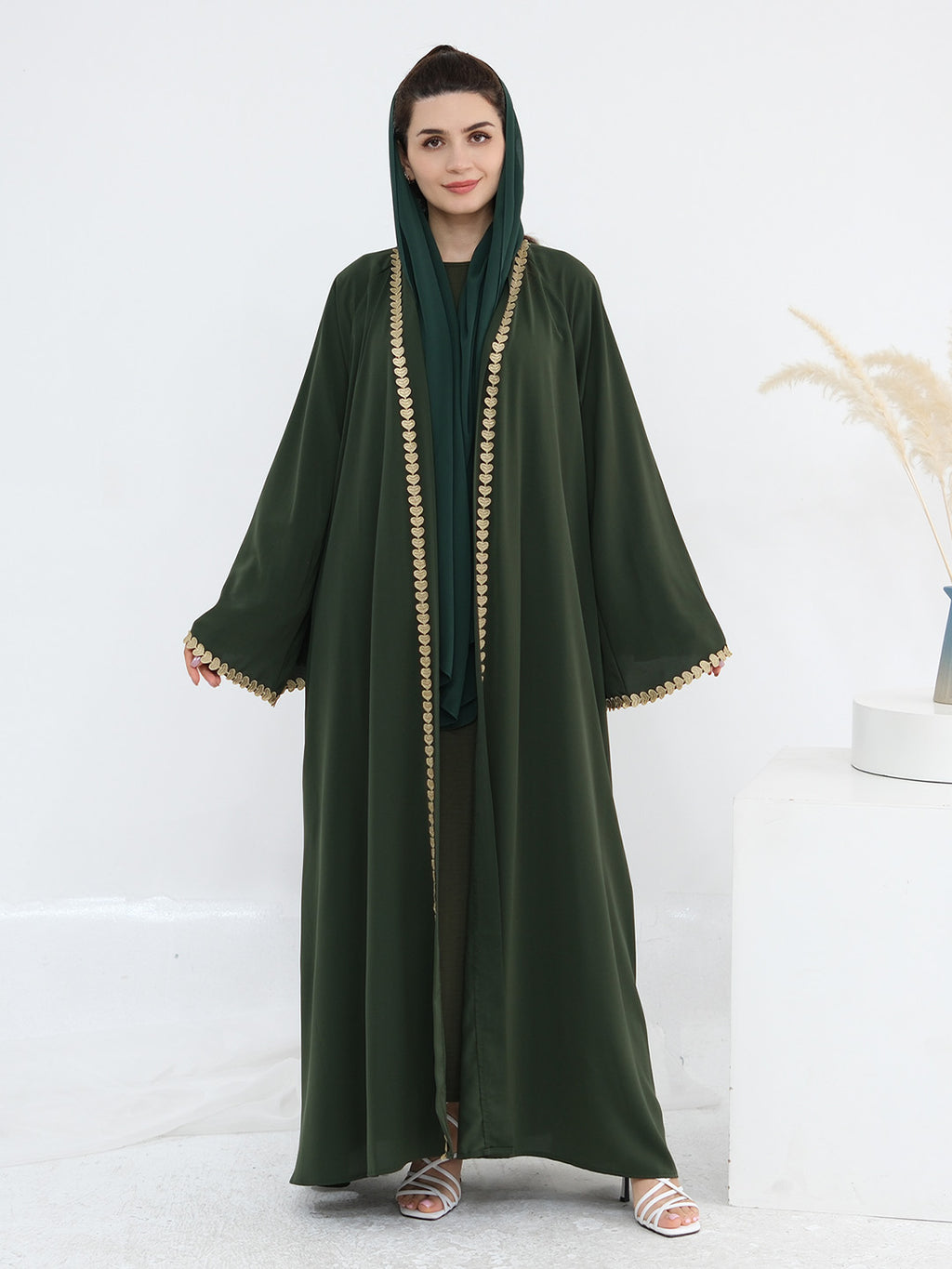 A woman modeling a dark green abaya with gold trim and a matching green headscarf, standing against a clean white studio background.
