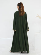 A woman viewed from behind wearing a long dark olive green abaya dress with wide sleeves and gold trim at the cuffs, standing in a bright white studio setting.