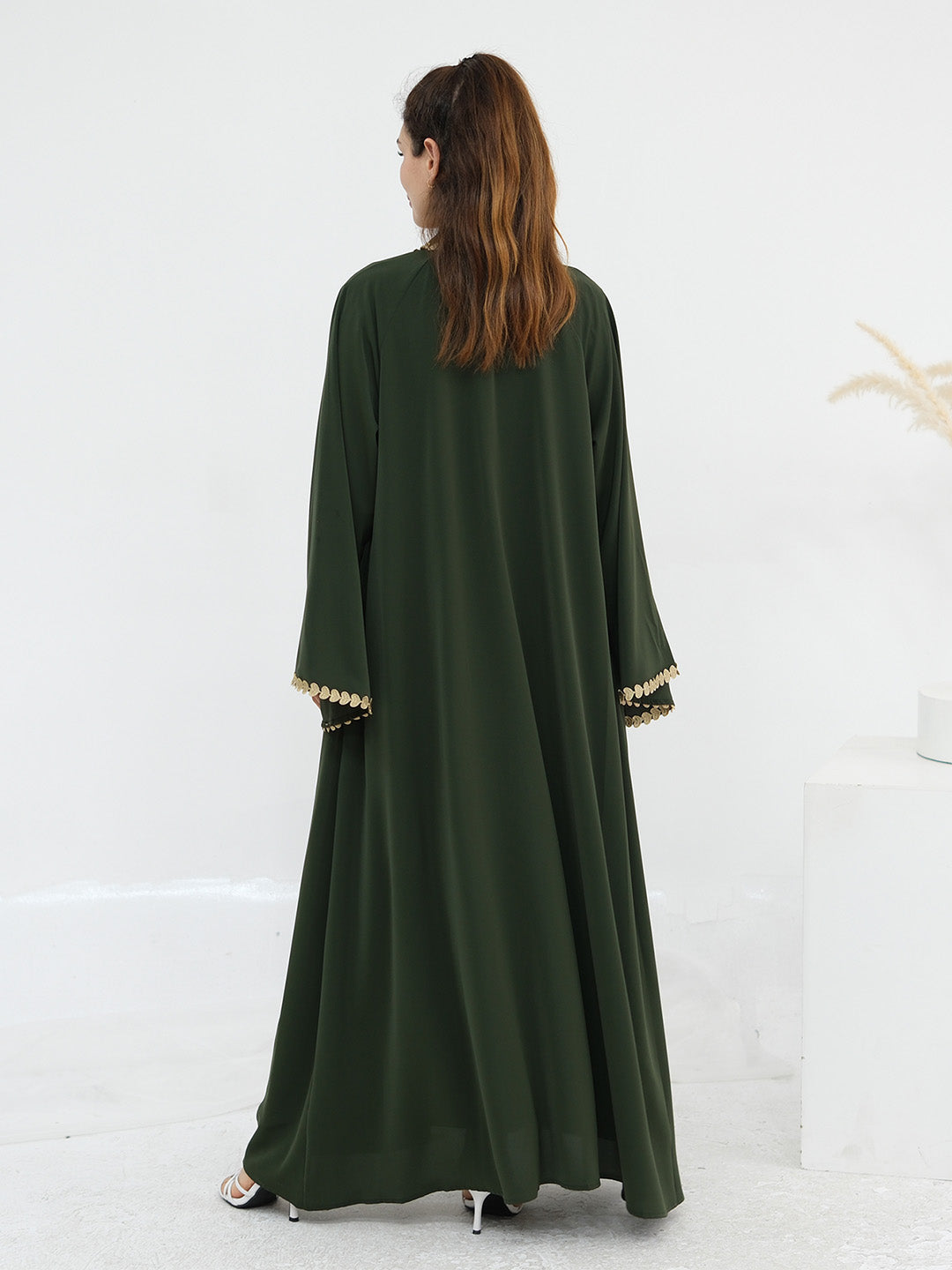 A woman viewed from behind wearing a long dark olive green abaya dress with wide sleeves and gold trim at the cuffs, standing in a bright white studio setting.