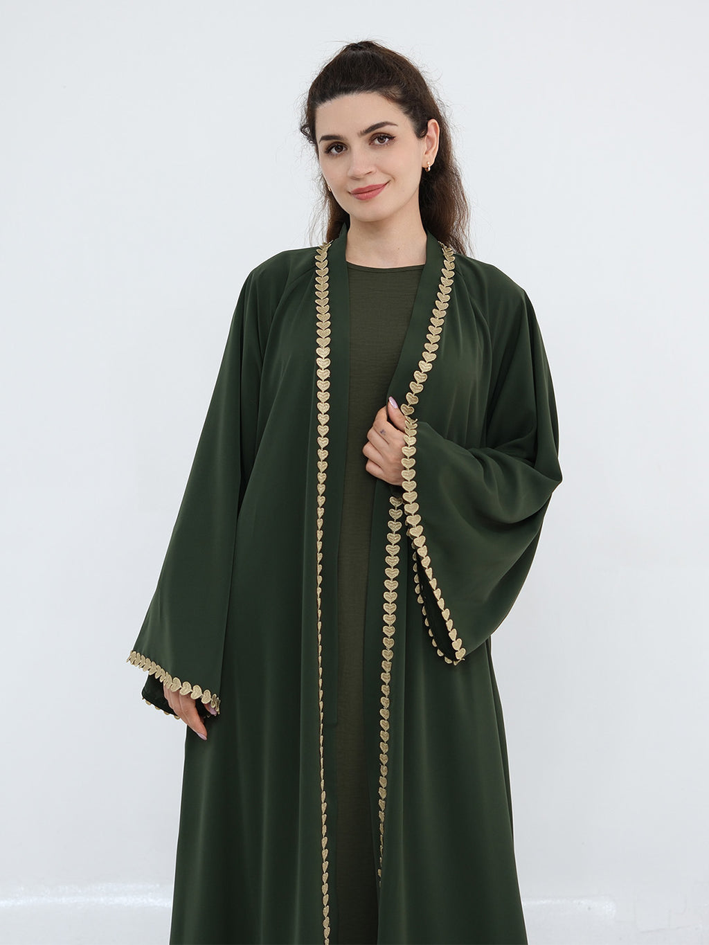 A woman modeling a dark green abaya with ornate gold coin trim along the front opening, sleeve edges, and cuffs, photographed against a plain light gray studio background.