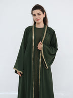 A woman modeling a dark green abaya with ornate gold coin trim along the front opening, sleeve edges, and cuffs, photographed against a plain light gray studio background.