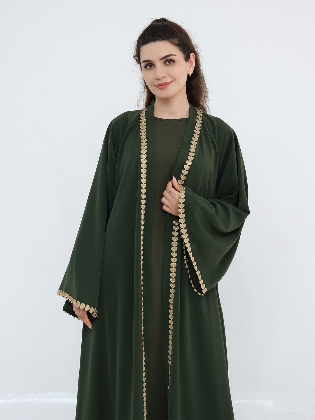 A woman modeling a dark green abaya with ornate gold coin trim along the front opening, sleeve edges, and cuffs, photographed against a plain light gray studio background.