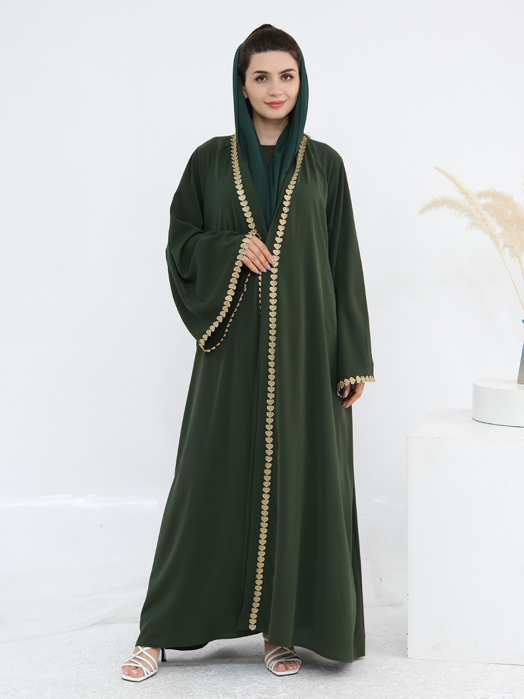 A woman modeling a dark green abaya with gold chain trim and a matching green hijab, standing against a clean white studio background.