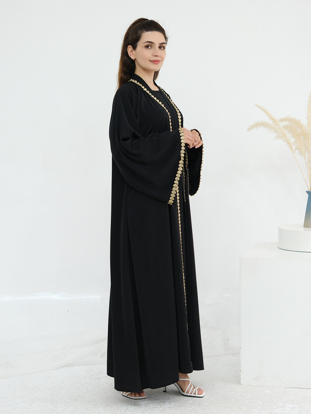 A woman modeling a long black abaya with gold chain detailing along the neckline, front opening, and side drape, standing in a bright white studio setting.