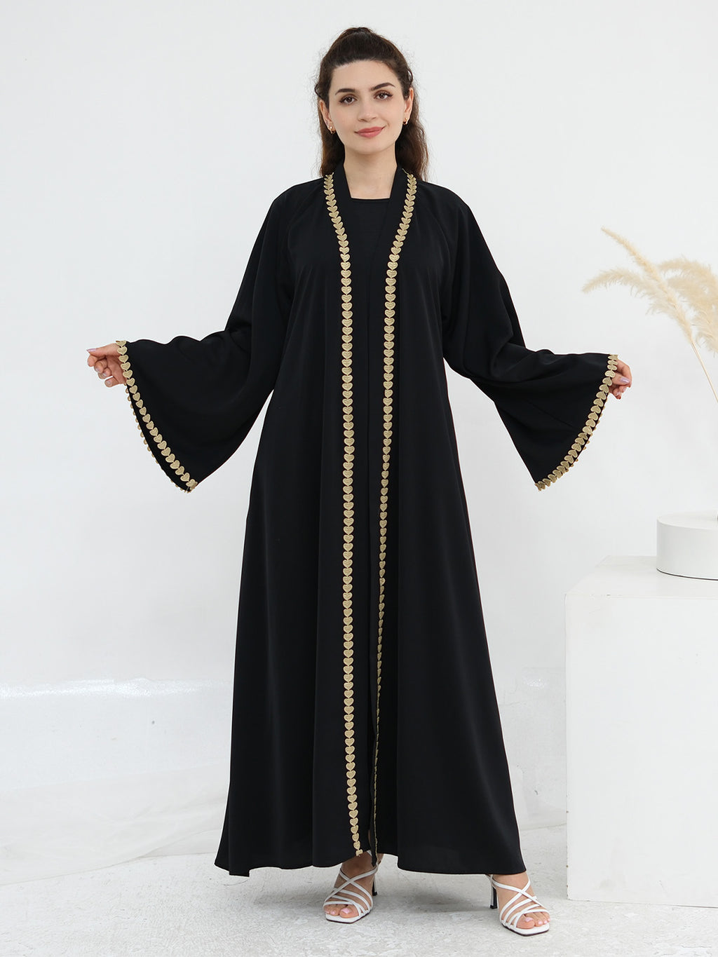 A woman modeling a long black abaya dress with wide bell sleeves and gold beaded trim along the front opening and sleeve cuffs, photographed against a clean white studio background.