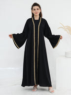 A woman modeling a long black abaya dress with wide bell sleeves and gold beaded trim along the front opening and sleeve cuffs, photographed against a clean white studio background.