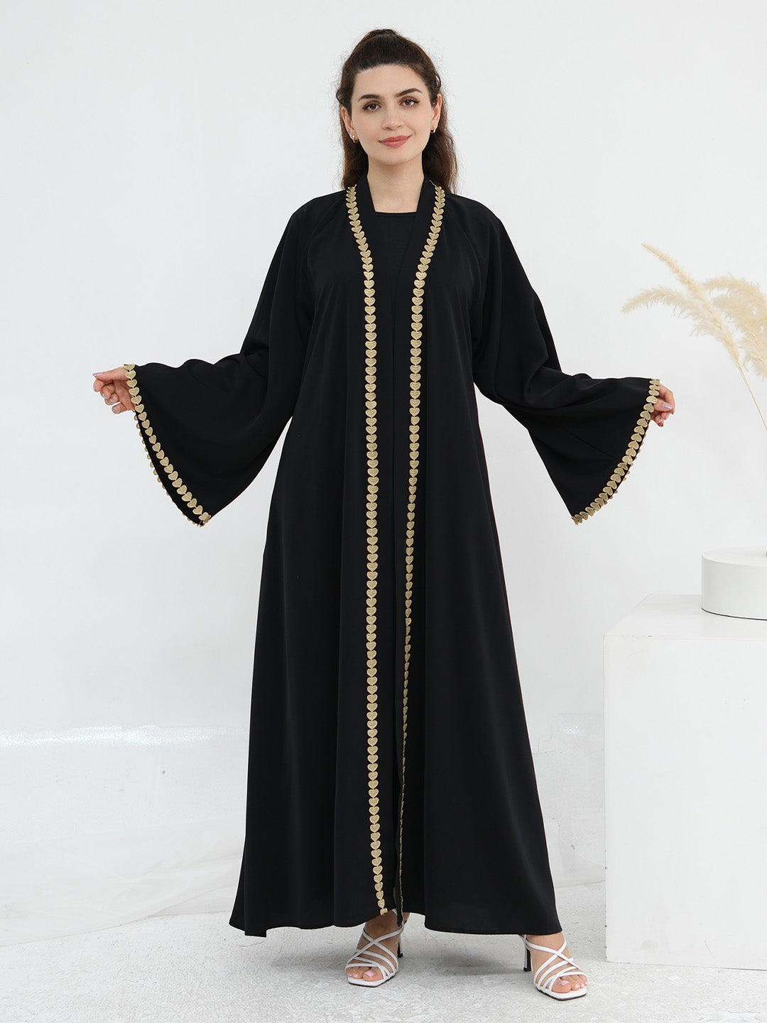 A woman modeling a long black abaya dress with wide bell sleeves and gold beaded trim along the front opening and sleeve cuffs, photographed against a clean white studio background.