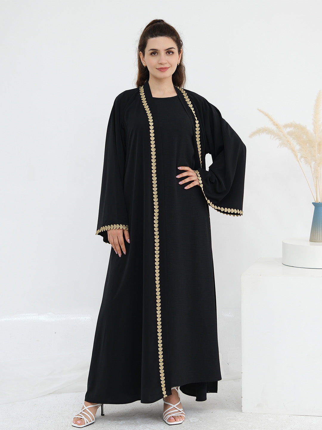 A woman modeling a long black abaya dress with ornate gold beaded trim along the front opening, sleeve edges, and hem, standing against a clean white studio background with a decorative pedestal and dried grasses.