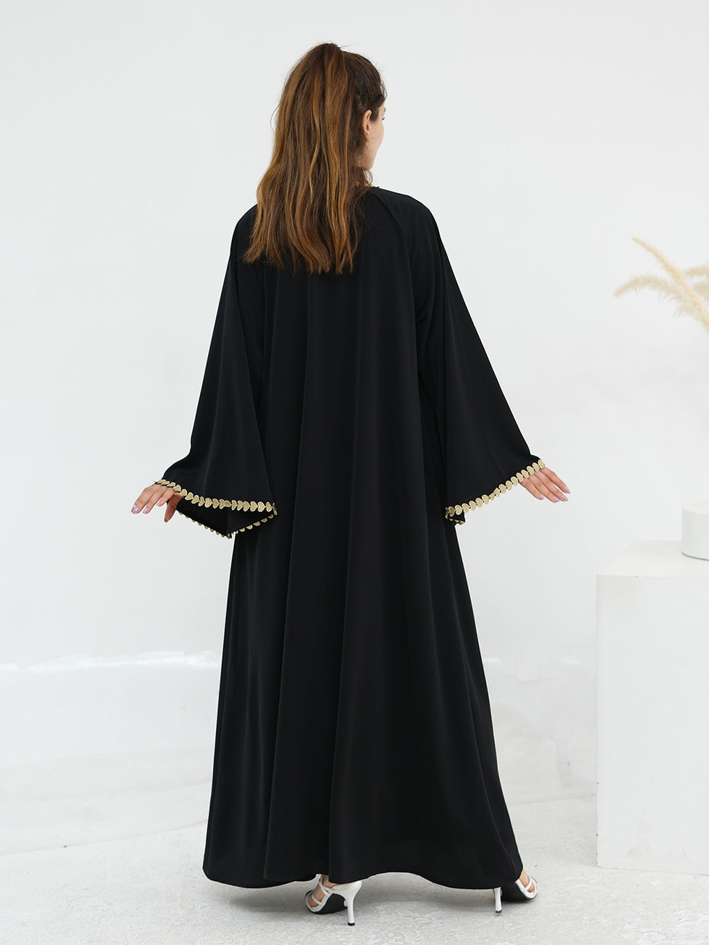 A woman viewed from behind wearing a long black abaya with wide sleeves and gold trim along the sleeve cuffs, standing in a bright white studio setting.