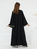 A woman viewed from behind wearing a long black abaya with wide sleeves and gold trim along the sleeve cuffs, standing in a bright white studio setting.