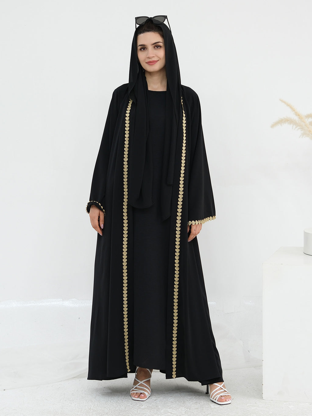 A woman modeling a black abaya with gold chain-like trim down the front and along the sleeve cuffs, styled with a matching black headscarf and sunglasses on her head, standing against a clean white studio background.