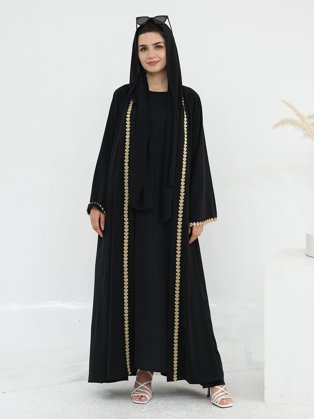 A woman modeling a black abaya with gold chain-like trim down the front and along the sleeve cuffs, styled with a matching black headscarf and sunglasses on her head, standing against a clean white studio background.
