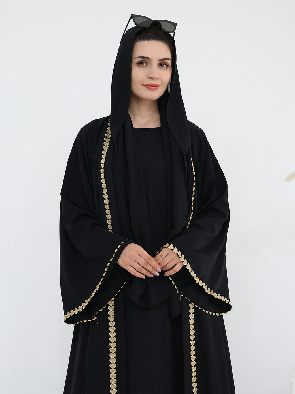 A woman wearing a black abaya with gold chain-like trim along the edges, a matching black head covering, and sunglasses on her head, photographed against a plain light background.