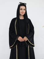 A woman wearing a black abaya with gold chain-like trim along the edges, a matching black head covering, and sunglasses on her head, photographed against a plain light background.