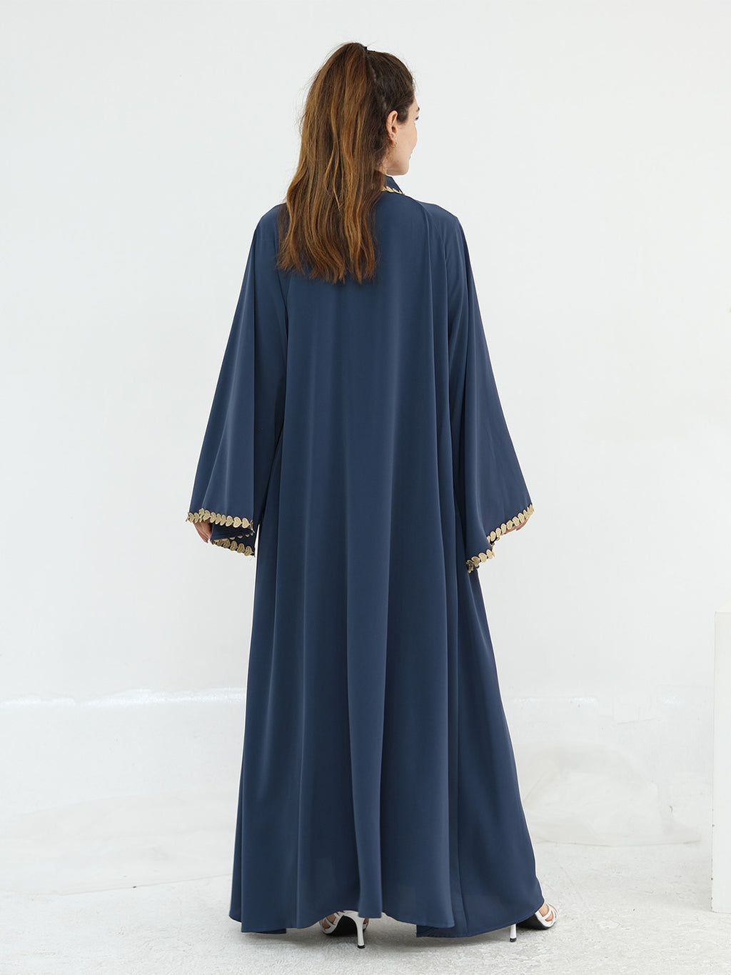 A woman viewed from behind wearing a long navy blue abaya with wide sleeves and gold trim at the cuffs, standing against a plain white studio background.