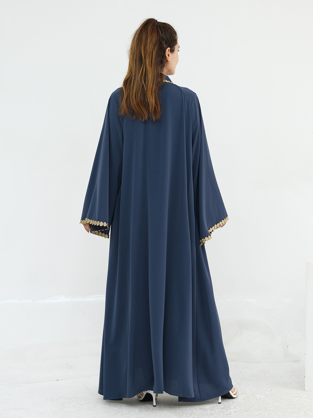A woman viewed from behind wearing a long navy blue abaya with wide sleeves and gold trim at the cuffs, standing against a plain white studio background.
