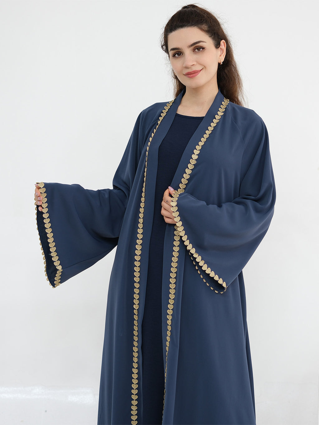 A woman modeling a long navy blue abaya with wide bell sleeves and a decorative gold coin border along the front opening and sleeve cuffs, set against a plain white studio background.