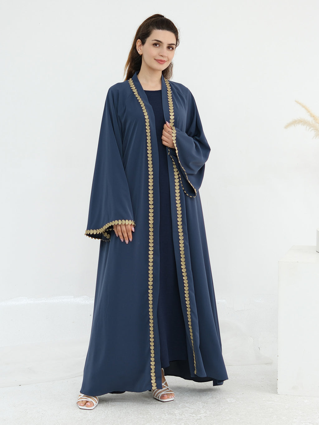 A woman modeling a navy blue abaya with ornate gold embroidery along the front opening, cuffs, and hem, set against a clean white studio background.
