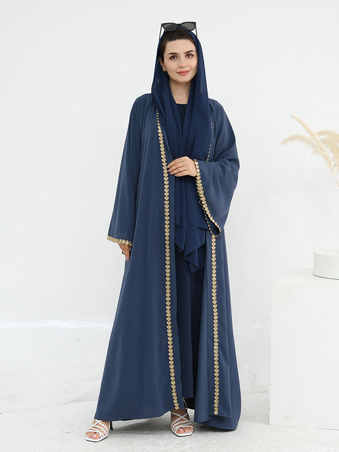 A woman modeling a navy blue abaya set with a matching headscarf, featuring gold embroidered trim down the front, on a clean white studio background.