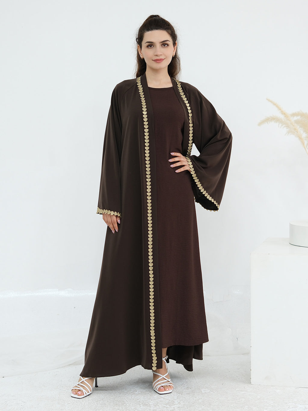 A woman modeling a long, dark brown abaya dress with wide sleeves and gold beaded trim along the front opening, cuffs, and hem, standing against a clean white studio background.