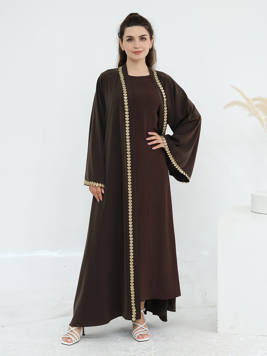 A woman modeling a long, dark brown abaya dress with wide sleeves and gold beaded trim along the front opening, cuffs, and hem, standing against a clean white studio background.