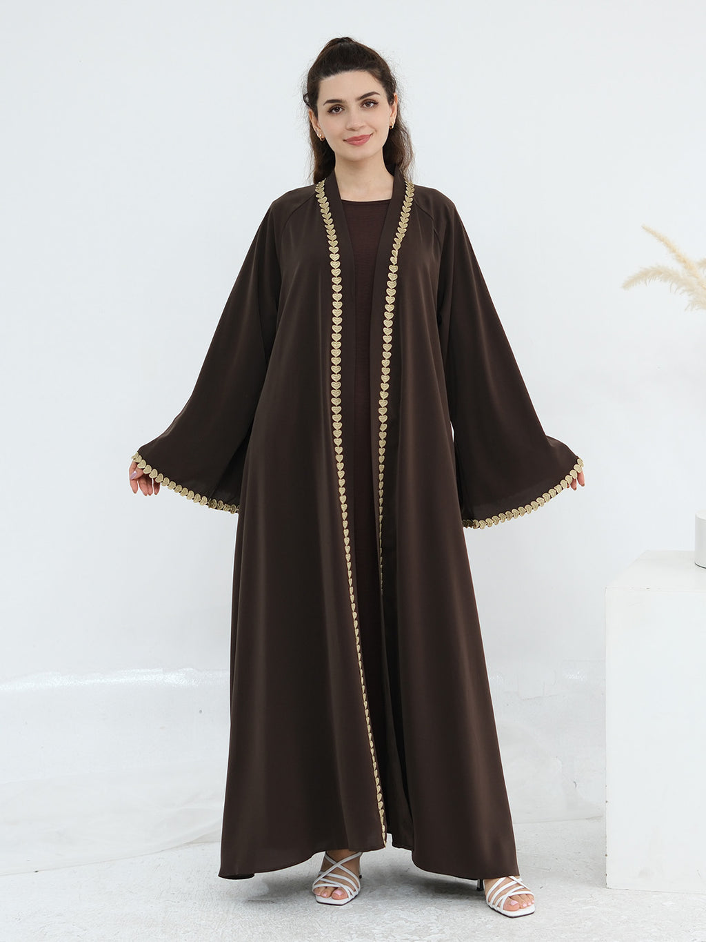 A woman modeling a dark brown abaya with wide sleeves and gold chain-like trim along the front opening and sleeve cuffs, standing against a clean white studio background.
