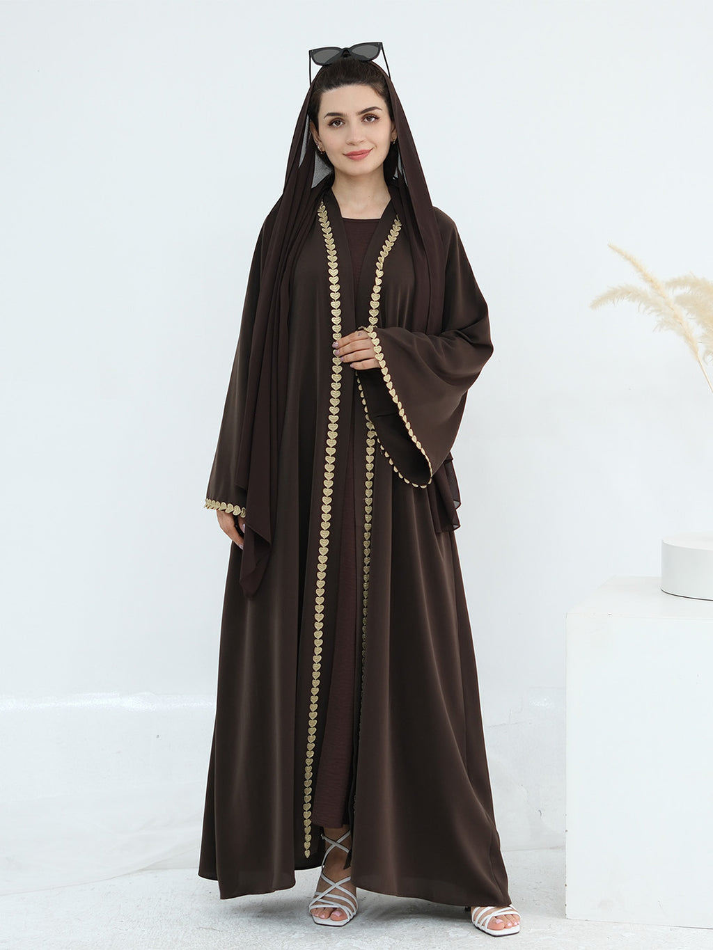 A woman modeling a dark brown abaya with a matching hood and long sleeves, featuring gold chain trim down the front and at the cuffs, paired with a matching inner dress and white heeled sandals.