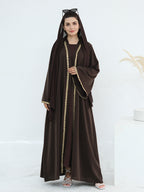 A woman modeling a dark brown abaya with a matching hood and long sleeves, featuring gold chain trim down the front and at the cuffs, paired with a matching inner dress and white heeled sandals.