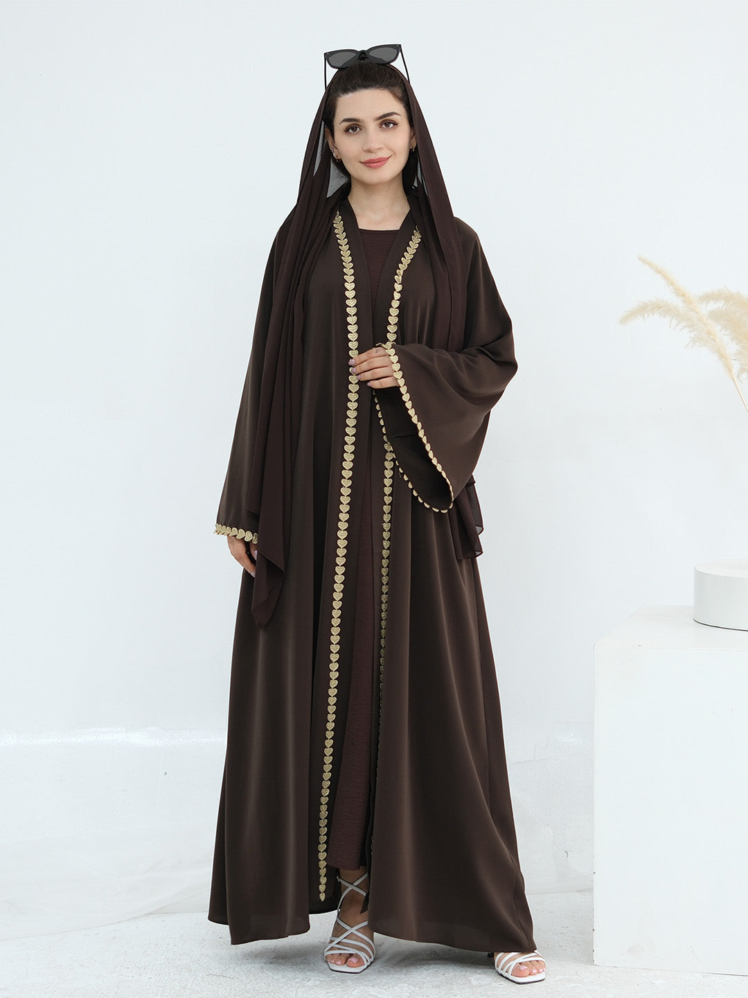 A woman modeling a dark brown abaya with a matching hood and long sleeves, featuring gold chain trim down the front and at the cuffs, paired with a matching inner dress and white heeled sandals.