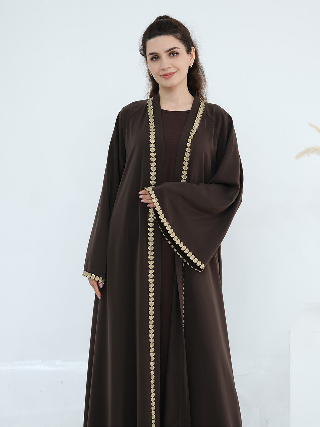 A woman modeling a dark brown abaya with ornate gold coin-like trim along the front opening, sleeve edges, and hem, set against a clean white studio background.