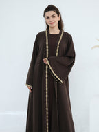 A woman modeling a dark brown abaya with ornate gold coin-like trim along the front opening, sleeve edges, and hem, set against a clean white studio background.