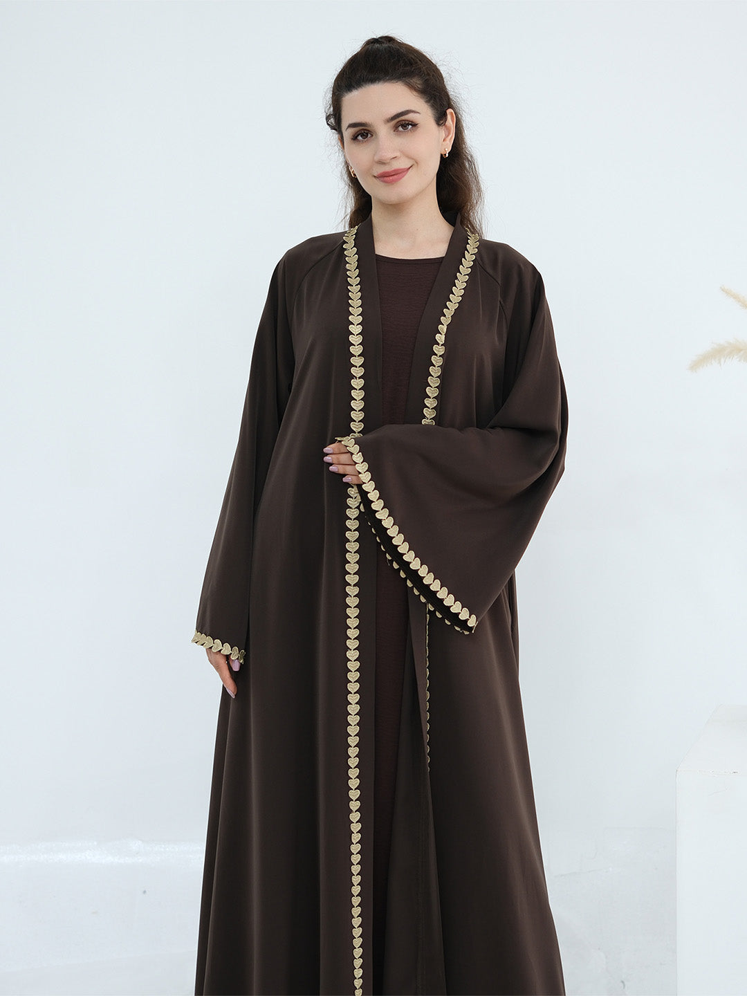 A woman modeling a dark brown abaya with ornate gold coin-like trim along the front opening, sleeve edges, and hem, set against a clean white studio background.