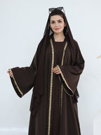 A woman wearing a dark brown abaya with wide bell sleeves and gold coin-trimmed edges, a matching head covering, and sunglasses resting on her head, set against a plain light background.