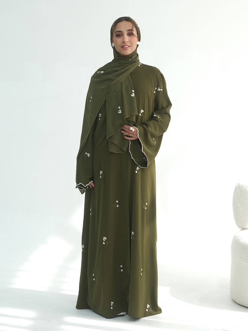 A woman wearing a long olive green abaya dress with a matching hijab, decorated with small white embroidered details, standing against a bright white studio background.