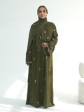A woman wearing a long olive green abaya dress with a matching hijab, decorated with small white embroidered details, standing against a bright white studio background.