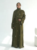 A woman wearing a long olive green abaya dress with a matching hijab, decorated with small white embroidered details, standing against a bright white studio background.