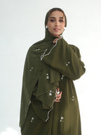 A woman wearing an olive green modest outfit with a matching headscarf, featuring delicate white floral embroidery and white piping details, set against a plain light gray background.