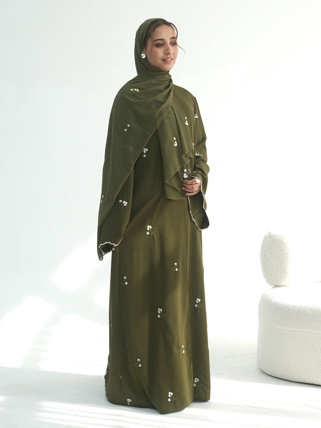 A woman modeling a full-length olive green abaya dress with a matching hijab, decorated with small white embroidered details, set against a bright white studio background with a modern white chair on the right.