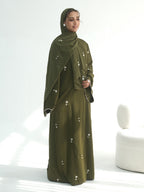 A woman modeling a full-length olive green abaya dress with a matching hijab, decorated with small white embroidered details, set against a bright white studio background with a modern white chair on the right.
