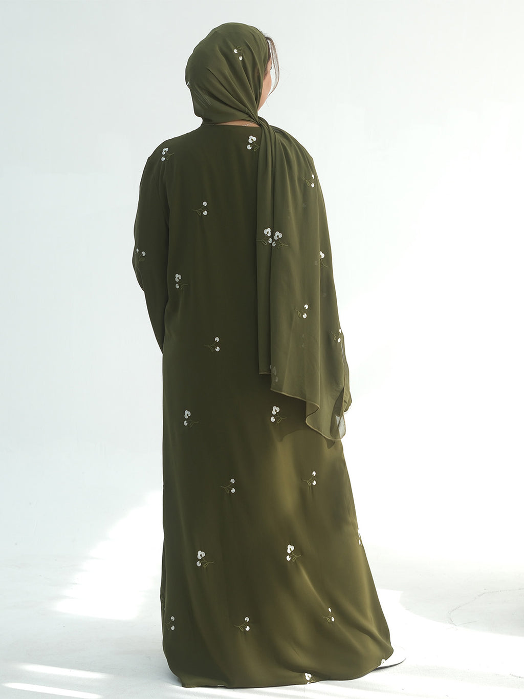 A woman viewed from behind wearing a long olive green abaya with small white embroidered details and a matching hijab, standing against a bright white studio background.