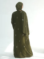 A woman viewed from behind wearing a long olive green abaya with small white embroidered details and a matching hijab, standing against a bright white studio background.