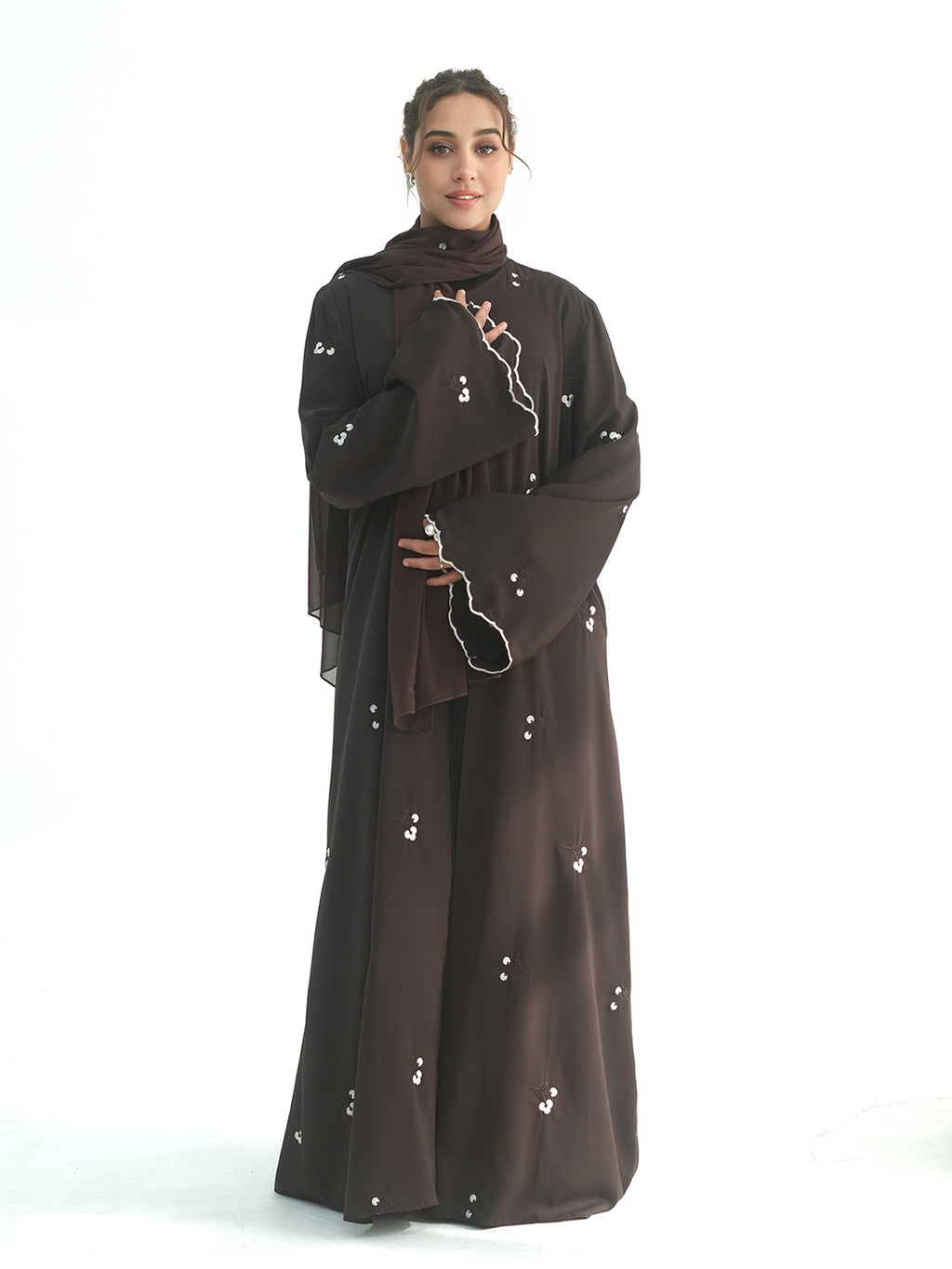 A woman modeling a dark brown abaya with matching hijab and wide sleeves, decorated with small white floral embroidery, set against a plain white background.